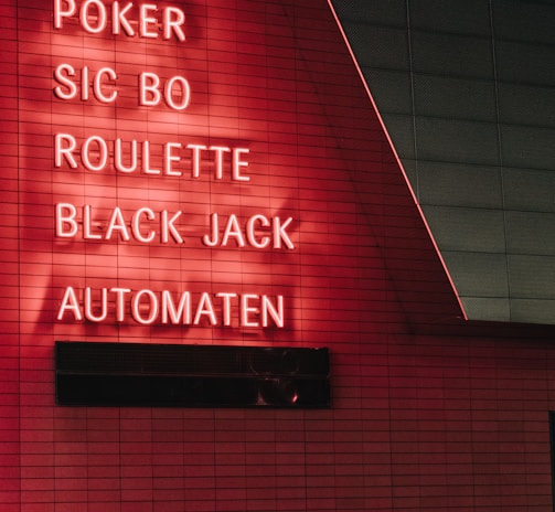 Red neon letters are mounted on a dark tiled wall, displaying the words Poker, Sic Bo, Roulette, Black Jack, and Automaten.