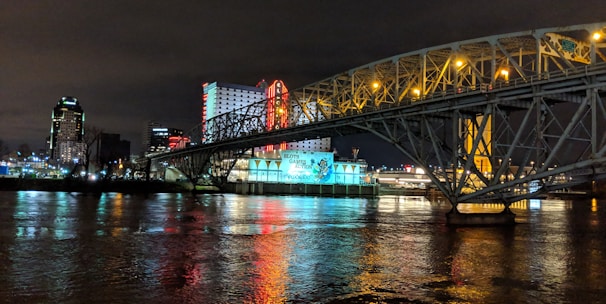 A brightly lit casino with neon signs and red lights is reflected in a river. A bridge stretches across the water, with an urban skyline visible in the background against an overcast night sky.
