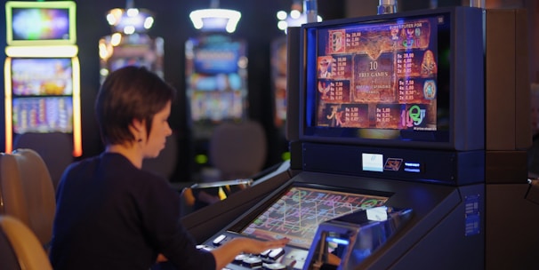 A person is seated at a slot machine console in a casino, focusing intently on the game displayed on the screen. The background shows several other illuminated gaming machines, adding a vibrant and colorful atmosphere to the scene.