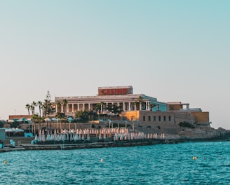 A seaside view of a large building labeled as a casino, situated on a rocky shore. The building is surrounded by palm trees and other smaller structures. The calm sea water reflects the sky, and there are several closed umbrellas lined along the waterfront.