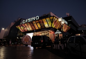 A building with a brightly illuminated entrance is seen at night. The sign above reads 'Casino' in bright letters. The facade features colorful graphics depicting various indulgent themes. Several vehicles are parked near the entrance, and people are visible in the vicinity. Tall lamp posts with spherical lights are present, casting a warm glow.