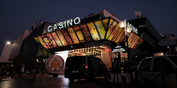 A building with a brightly illuminated entrance is seen at night. The sign above reads 'Casino' in bright letters. The facade features colorful graphics depicting various indulgent themes. Several vehicles are parked near the entrance, and people are visible in the vicinity. Tall lamp posts with spherical lights are present, casting a warm glow.