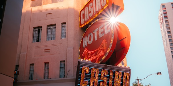 A vibrant city scene featuring a tall, illuminated casino sign with retro-style neon lights. The sign dominates the building facade with the words 'CASINO' and 'HOTEL'. Sunlight peeks through, creating a dramatic lens flare effect. The street is lively, with pedestrians casually walking and a hint of urban hustle.